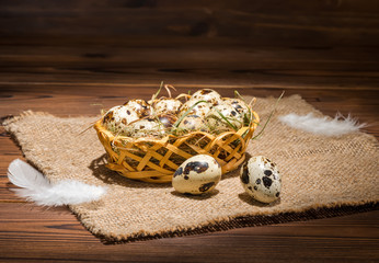 quail eggs with straw and feathers in basket on sackcloth wooden background, rustic style, close up