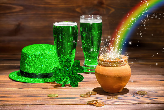 St Patricks day concept with glasses of green beer, shamrock, leprechaun hat, pot full gold coins, spilling coins and rainbow on vintage wooden background, close up
