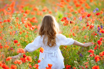  young girl wearing white dress in summer blooming field walking under evening sun. View from back