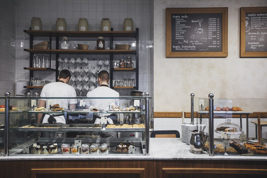 Two young men working in coffee shop at bar with pastry showcase.
