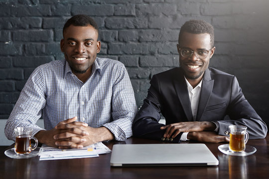 Portrait Of Two Cheerful African-American Businessmen Or Business Partners Sitting At Desk With Laptop Computer And Papers, Ready To Start Meeting, Both Looking At Camera With Happy Expression