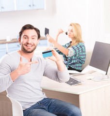 Toned of happy young man smiling for camera and showing credit card. Bearded man sitting at table with computer on while his girl friend powdering her face on background.