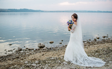 Brunette bride with violet wedding bouquet near beach of river at sunset.