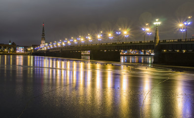 Fototapeta na wymiar illuminated stone bridge Akmens tilts leading over the frozen river Daugava to the St. Peter's Church in the old part of the city of Riga, Latvia on a cold winter evening