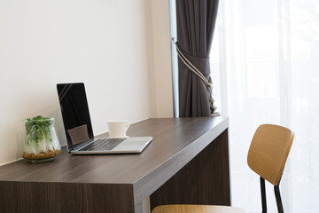 office table with blank screen laptop, white coffee cup, modern chair and garden plant garden on cement wall background.