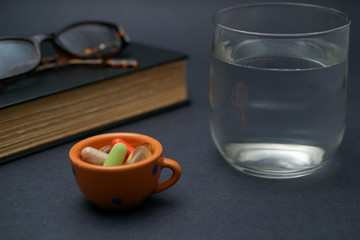 A pair of broken reader glasses resting on an old open book,  a bunch of pills in a bowl and a cup of water on black background, suggesting an elderly person taking a break take medication