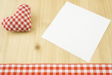 Ein rot weiß kariertes Herz mit weißem Zettel und kariertem Band am Bildunterrand, Holzhintergrund, Textfreiraum