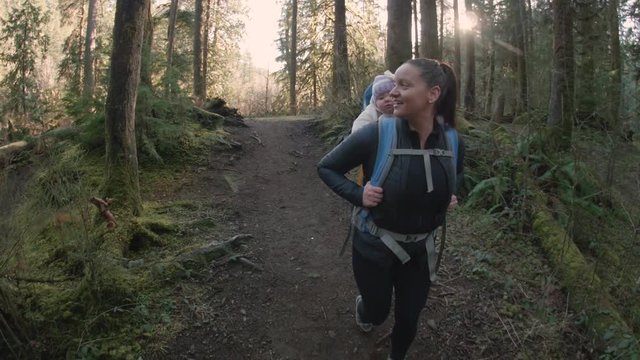 Happy Mom and Baby Daughter in Hiking Backpack Walking Down Forest Trail on Sunny Day