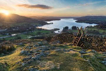 Fototapeta premium Sun rising above the horizon illuminating the landscape with stone wall and stile in foreground.