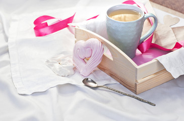 A tray with a cup of hot tea and homemade meringue in the form of heart in a bed on a white textile