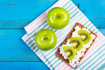 Useful snack, quick healthy meal. Grain bread with homemade curd cheese and kiwi slices on a blue wooden background. Two juicy green apples on the table napkins.