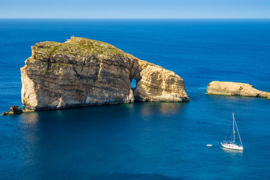 Gozo, Malta - The amazing Fungus Rock at Dwejra bay with sailboat, blue sea water and sky on a beautiful summer day