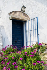 Fototapeta na wymiar Spanish blue old entrance door with the open gate in white house, purple chrysantemum flowers in front
