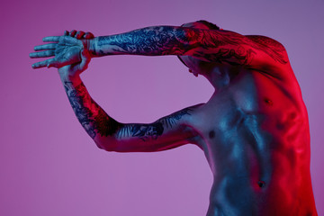 Fashion photoshoot of sport fit attractive man making arm stretch. Masculine naked body, tattooed hands, hipster look. Color flash studio light