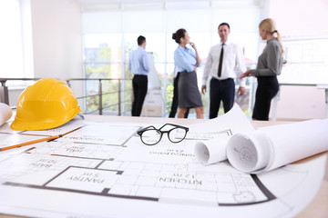 Helmet with blueprints and glasses on table and engineers on blurred background