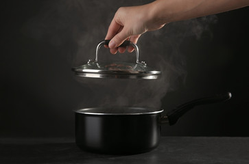 Human hand removing cover from saucepan with hot liquid, on dark background