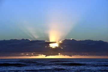 Beautiful sunrise cloudscape over ocean background. Sun rays beaming through picturesque clouds above sea. Blue sky with clouds, sea and sun on the horizon. Beautiful sunrise over the quiet calm sea.