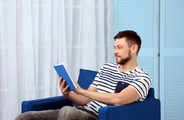 Handsome young man reading book while sitting in arm-chair at home