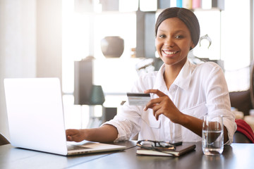 Good looking young black woman smiling at the camera while seated in front of her computer at her dining room table behind her notebook where she is making online payments with her credit card.