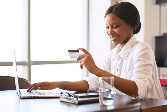 Young Black Woman Smiling Happily While Making Electronic Payments With The Use Of Her Credit Card That She Is Holding In One Hand While Typing With The Other.