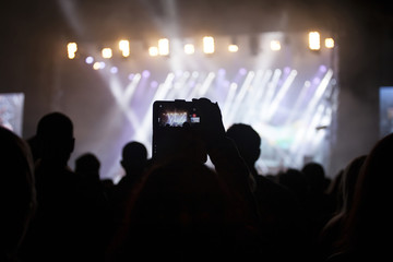 Close up on hands taking photos with phone on a concert, colorful stage in the background.