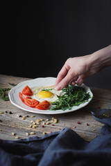 Breakfast with fried egg, green salad and fresh tomatoes. Female hand decorating plate with breakfast. Selective focus, rustic style