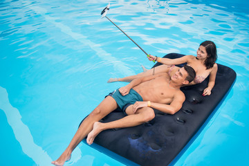 Smiling couple floating on mattress in pool and taking selfie photo on the phone with selfie stick on their summer vacation. Copy space