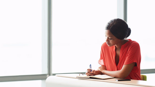 Adult Female African Writer Busy Writing In Her Notebook While Wearing A Bright Orange Blouse While Seated Next To Large Panes Of Glass In The Business Lounge She Is Seated In.