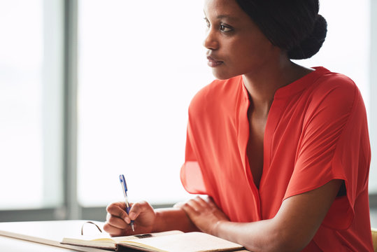 Black Female Creative Writer Busy Looking Up Into The Distance As She Procrastinates And Takes A Break From Her Writing Session While Dressed In A Colourful Orange Shirt.