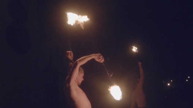 Fire show street circus performance close up slow motion. Two male fire performers spinning burning fire poi at black background in night during carnival