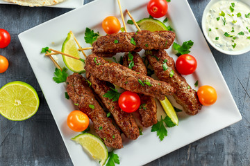 Homemade Kofta kebabs on skewers with pita, lime, vegetables, sweet chili and yogurt sauce on white plate.