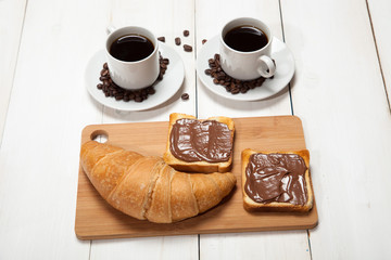 Cups of coffee with a croissant and toast with chocolate on a white wooden table