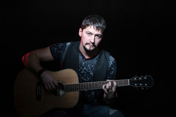 Obraz na płótnie Canvas Portrait of handsome bearded man sitting in a chair with an acoustic guitar in hand, isolated on a black background