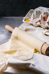 Fresh homemade rolled dough for pasta tagliatelle on wood rolling pin with egg yolk and white linen textile over dark gray table background.