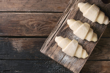 Fototapeta na wymiar Raw unbaked croissant on wooden chopping board over old dark wood background. Top view, copy space