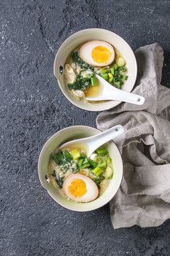 Two bowls with asian style soup with scrambled eggs, half of marinated egg, spring onion, spinach served with white spoons and textile over black texture concrete background. Top view with space