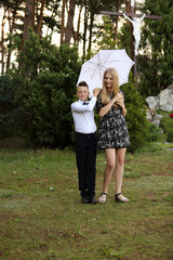 Siostra i brat stoją uśmiechnięci pod parasolem w parku. - obrazy, fototapety, plakaty