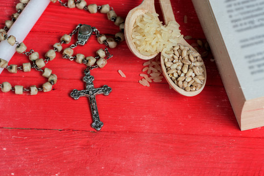 silver Christian cross necklace on white candle,  Bible and seeds of rice and wheat in a wooden spoon - bright red on the old table. as a symbol of the beginning of Great Lent - Toned image  