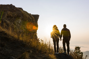Back view of young couple hikers standing on top of the mountain enjoying sunrise over the tropical valley. ,Happy couple holding hands looking in the sunset.,Romantic life moment at warm orange light