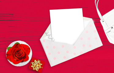     White woman things on the red wooden background. Red rose in vase, white envelope, white woman bag 