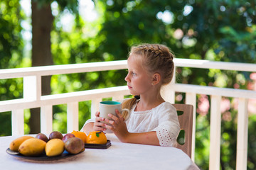 Cute beautiful girl drinks morning for breakfast vitamin juice. Girl thoughtfully looking off the balcony on the palm grove. On the table next to pineapple and mango fruits.