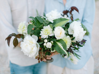 the man and woman dressed in light clothes stand nearby and hold in hand beautiful flower composition from white colors outdoors.
