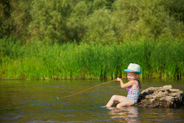 Cute little girl in funny hat pretends to catch fish, child plays fishing on river