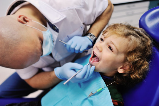 Male Dentist Examines The Teeth Of The Patient Cheerful Child With Curly Red Hair. Moloi Boy Smiling In Dentist's Chair. Child Mouth Wide Open In The Dentist's Chair