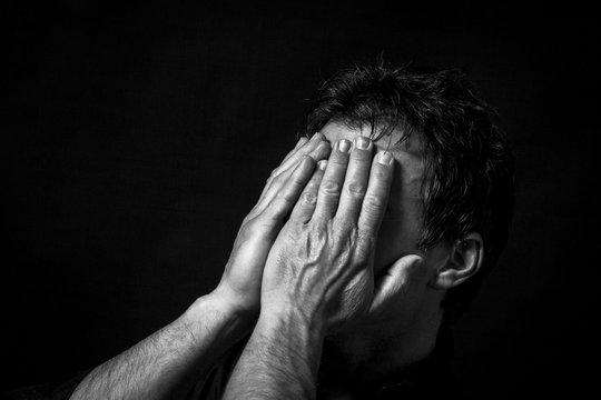Close up portrait of isolated desperated scared man, with hands on the face. Monochrome picture, black background
