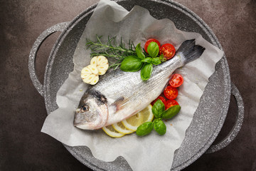 Fresh uncooked dorado or sea bream fish with lemon, herbs, oil, vegetables and spices in a frying pan on a baking paper over black backdrop, top view