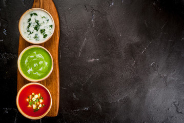 Selection of cold refreshing summer soups: tomato gazpacho, tarator, cream soup of green peas on a cutting board on a black concrete table top view copy space