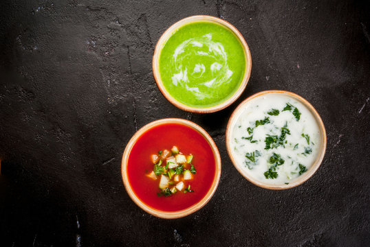 Selection of cold refreshing summer soups: tomato gazpacho, tarator, cream soup of green peas on a black concrete table top view copy space