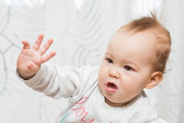 Eleven months old baby girl sitting in a stool making funny faces into the camera and raising her right arm in the air as if trying to stop someone or reach for something