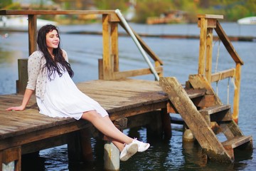 Adorable smiling long-haired brunette girl in a white dress and beige knitted cardigan sitting on wooden dock over the water with my eyes closed and enjoying warm spring Sunny day, side view.